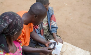 A male para social worker in an orange T-shirt sits between a mother and son, holding a sheet of paper with notes and a small clock.