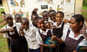 A group of school children in uniforms smile and wrap their arms around each other in front of a mural outside.