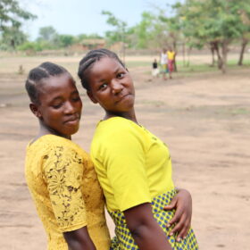 Two young women dressed in yellow stand side by side, leaning and smiling, against an open rural background.