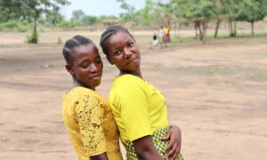 Two young women dressed in yellow stand side by side, leaning and smiling, against an open rural background.