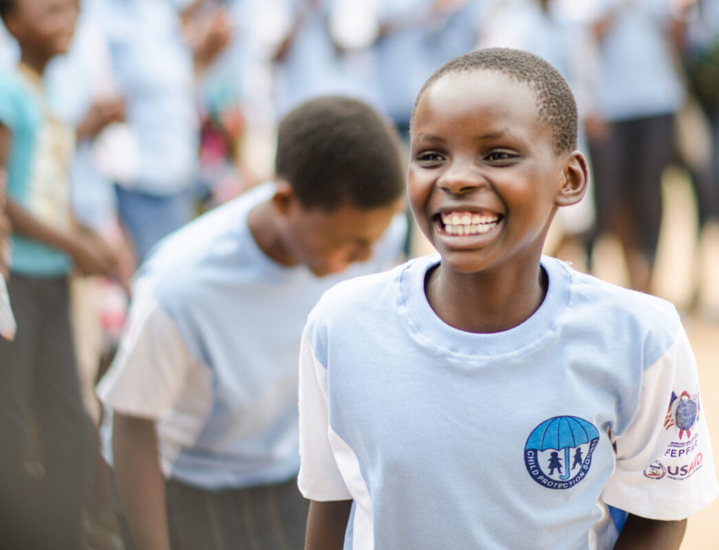 Young boy in focus flashes a big smile amongst a crowd of schoolmates, wearing a blue Child Protection Society T-shirt.