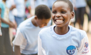 Young boy in focus flashes a big smile amongst a crowd of schoolmates, wearing a blue Child Protection Society T-shirt.