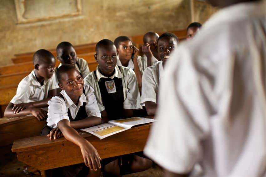 A group of children in school uniforms sit next to each other in a classroom looking at a teacher.