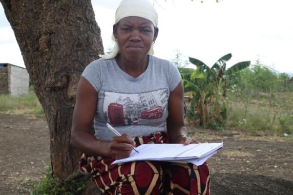 A women sitting at the base of a tree looks directly at the camera while taking notes on a notepad.